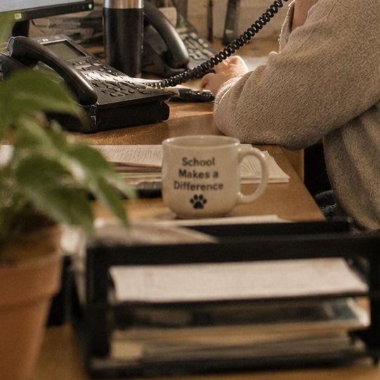 A school front-office desk: two phones, paperwork, and a mug that reads School Makes a Difference